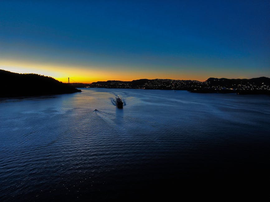 Aerial view of a ship sailing through a Norwegian fjord at sunset, showcasing vibrant twilight colors and serene waters.