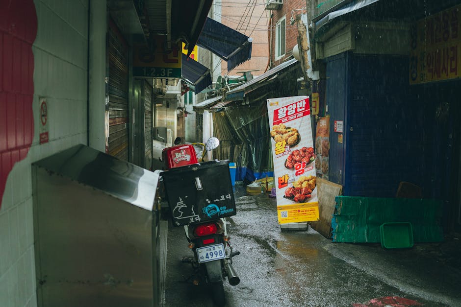A rainy day scene in a Seoul alleyway featuring a motorbike and food sign in a traditional market area.